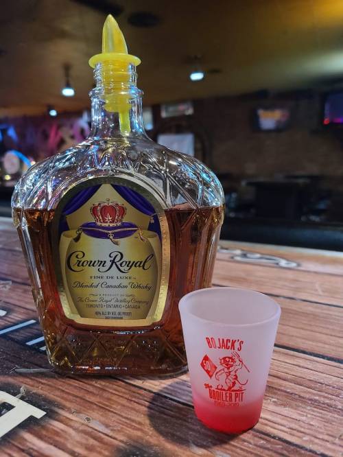 A bottle of Crown Royal whiskey next to a red shot glass on a wooden table in a bar setting.