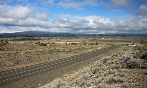 A winding road leads through a vast landscape of fields and mountains under a cloudy sky.