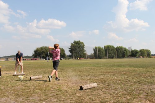A boy swings a bat at a log in a grassy field, while another person prepares to hit. Trees and a blue sky are in the background.