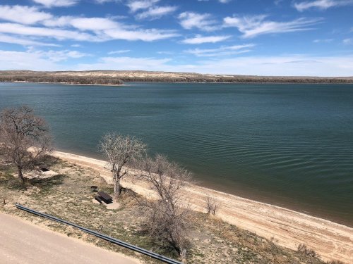 A serene lake view with sandy shores, trees, and blue skies, reflecting calm waters under a few clouds.