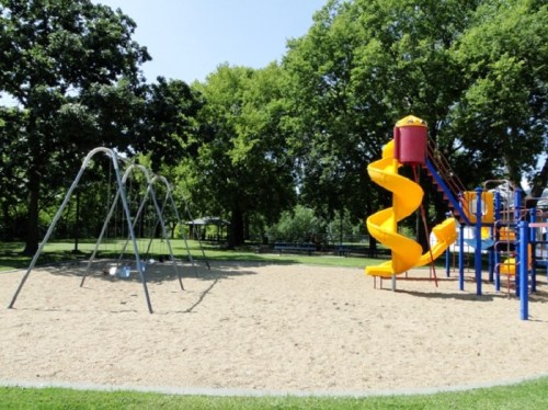 A sunny playground featuring a yellow slide, swings, and surrounded by green trees and sandy ground.