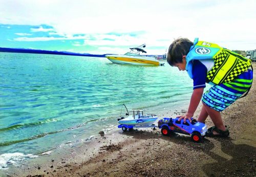 A child in a life jacket plays with toy boats and a truck on the shore of a lake, with a boat in the background.