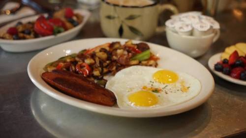 A plate with two sunny-side-up eggs, hash browns, and a slice of sausage, with fruit and coffee in the background.