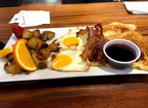 A breakfast plate with two eggs, bacon, toast, roasted potatoes, and a slice of orange, served with syrup.