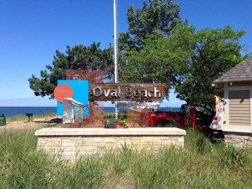 Sign for Oval Beach with a blue sky, trees, and a glimpse of the water in the background.