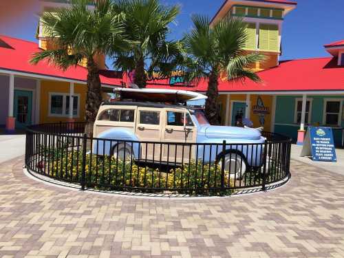 A vintage blue station wagon with a surfboard on top, surrounded by palm trees and colorful buildings.