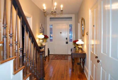 A well-lit hallway with a staircase, decorative lamps, and a door leading outside, featuring a rug and wall decor.