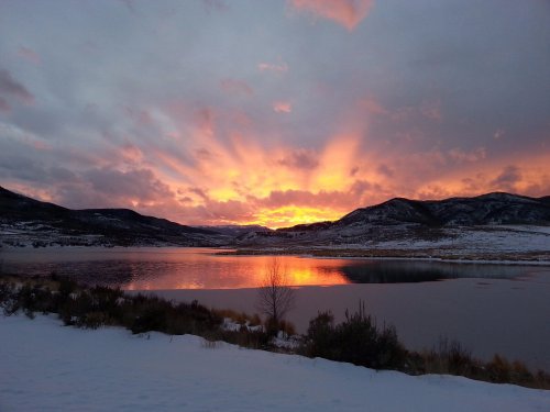 A vibrant sunset casts orange and pink hues over a snowy landscape and calm lake, framed by distant mountains.
