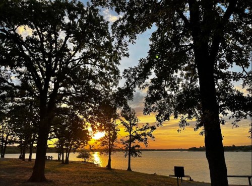 A serene lakeside scene at sunset, framed by trees, with vibrant colors reflecting on the water.