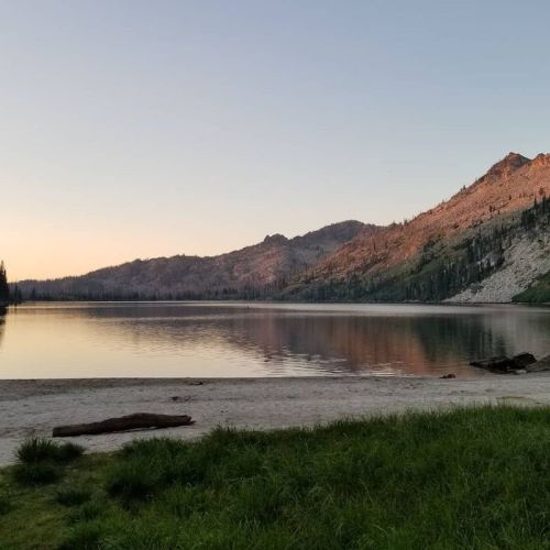 A serene lake at sunset, surrounded by mountains and trees, with a sandy shore and calm water reflecting the landscape.