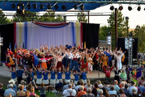 A large group of performers in colorful costumes celebrates on stage, while an audience watches from below.