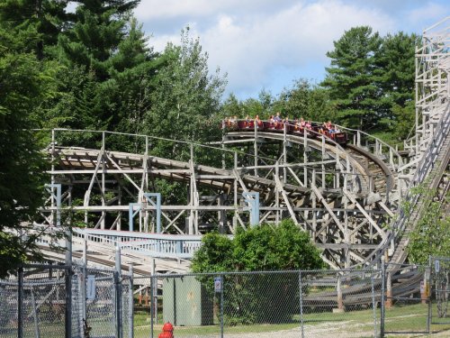 A wooden roller coaster with riders ascending a hill, surrounded by trees and a fence.