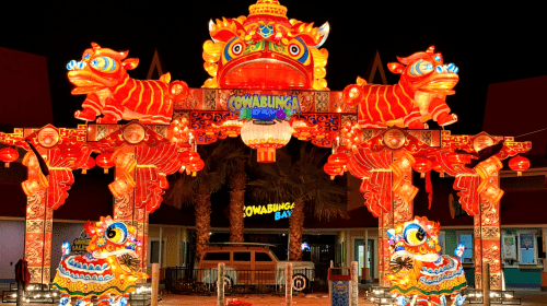 Colorful illuminated entrance with dragon sculptures at a festive night event, surrounded by palm trees.