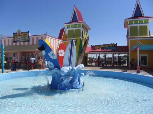 Colorful surfboards emerge from a fountain in a vibrant amusement park, with bright buildings in the background.