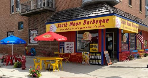 Colorful outdoor seating at South Moon Barbeque, featuring umbrellas and a welcoming sign on a brick building.