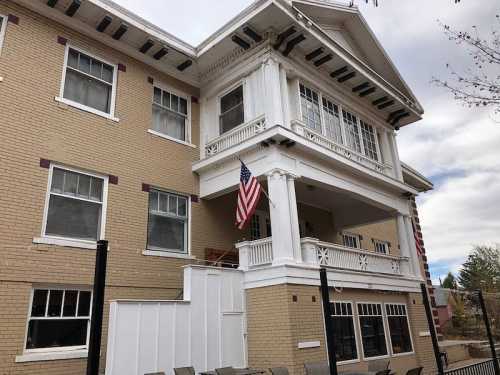 Historic building with a white porch, large windows, and an American flag, set against a cloudy sky.