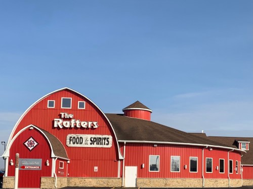 A red barn-style building with a round roof, featuring a sign that reads "The Rafters Food & Spirits." Clear blue sky.
