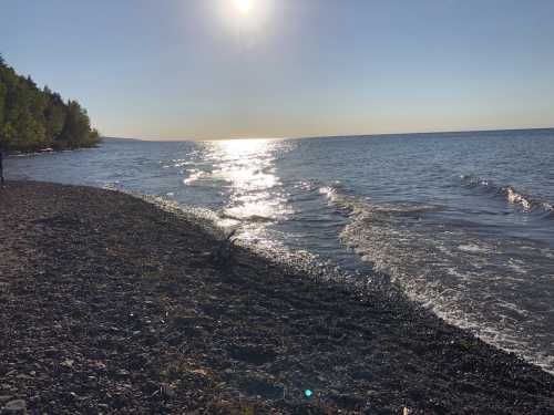 A serene beach scene with gentle waves, pebbles, and sunlight reflecting on the water under a clear blue sky.