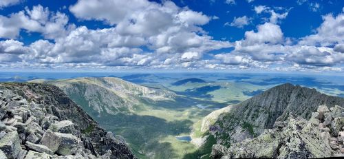 Panoramic view of rugged mountains and valleys under a bright blue sky with fluffy clouds.
