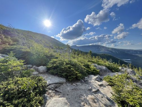 A scenic mountain landscape with lush greenery, rocky terrain, and a bright sun shining in a blue sky with clouds.