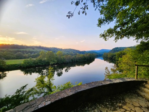 A serene river reflects the sky, surrounded by lush greenery and rolling hills at sunset.