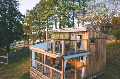 A wooden two-story cabin with a balcony, surrounded by trees, and a person standing on the upper level.
