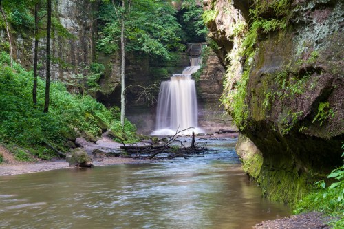 A serene waterfall cascades into a calm river, surrounded by lush greenery and rocky cliffs.