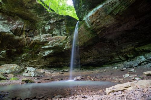 A serene waterfall cascades into a rocky pool beneath a moss-covered overhang in a lush green forest.