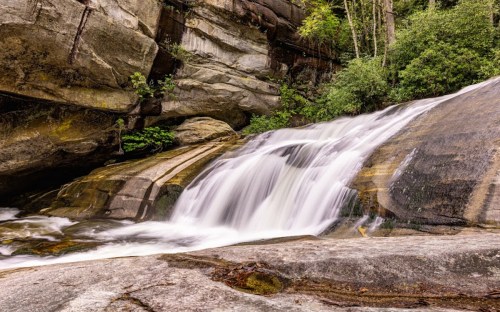 A serene waterfall cascading over smooth rocks, surrounded by lush greenery and forested cliffs.