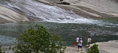 A family walks along a rocky shore near a flowing river and waterfall, surrounded by greenery.