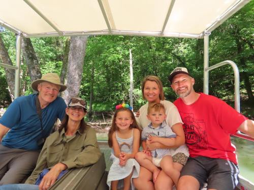 A group of six people, including children, smiling together on a boat surrounded by trees.