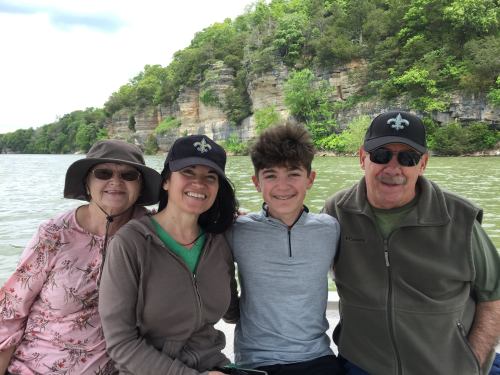 A family of four smiles together on a boat, surrounded by green cliffs and water on a sunny day.
