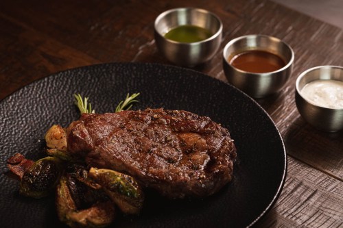 A beautifully plated steak with roasted Brussels sprouts, garnished with herbs, and accompanied by three dipping sauces.