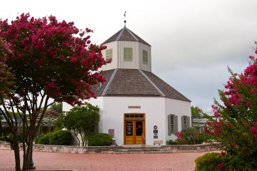 A charming white building with a gray roof, surrounded by vibrant pink flowering trees and a brick pathway.