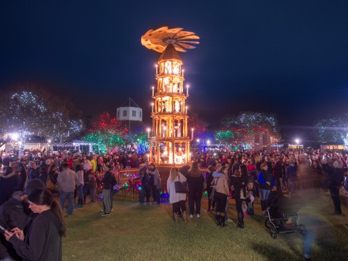 A festive crowd gathers around a tall, illuminated carousel at night, surrounded by colorful holiday lights.