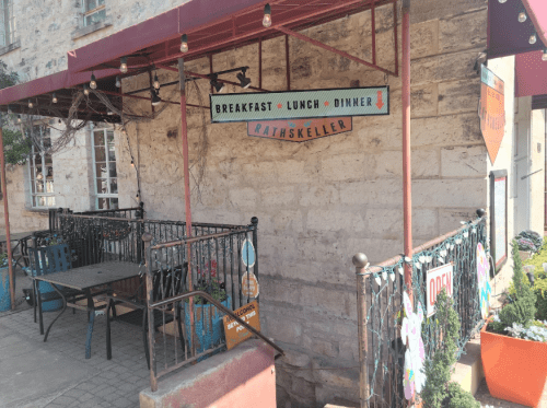 A cozy restaurant entrance with a sign for breakfast, lunch, and dinner, featuring colorful decorations and outdoor seating.