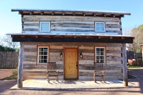 A rustic log cabin with a wooden door, two rocking chairs on the porch, and windows on the upper and lower levels.
