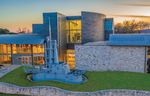 Aerial view of a modern building with a submarine sculpture in front, surrounded by green grass at sunset.