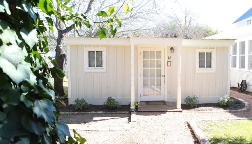 A small, light-colored cottage with a front porch and two windows, surrounded by greenery and a gravel path.