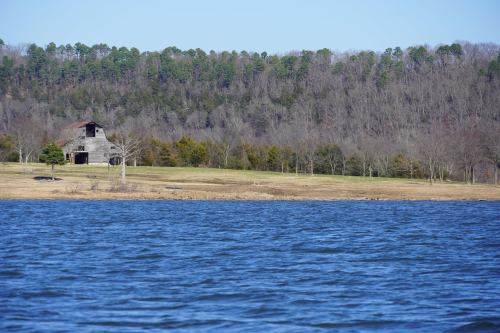 A serene lake view with a rustic barn and trees lining the shore under a clear blue sky.
