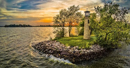 A serene lighthouse on a rocky island at sunset, surrounded by calm waters and lush greenery.