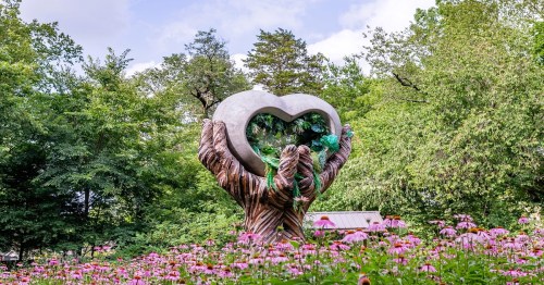 A large heart-shaped sculpture held by two hands, surrounded by colorful flowers and lush greenery.