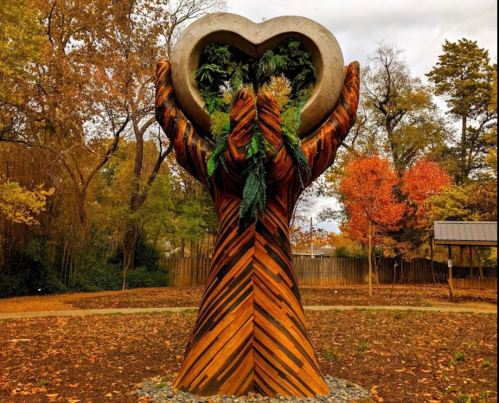 A wooden sculpture of hands holding a heart, adorned with greenery, set in a colorful autumn landscape.