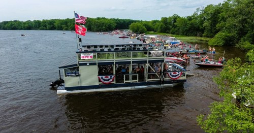 A floating restaurant on a river, surrounded by boats and greenery, with flags and festive decorations.