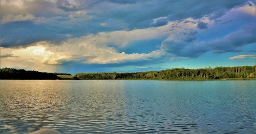 A serene lake scene with calm waters, lush green trees, and a dramatic sky filled with clouds at sunset.