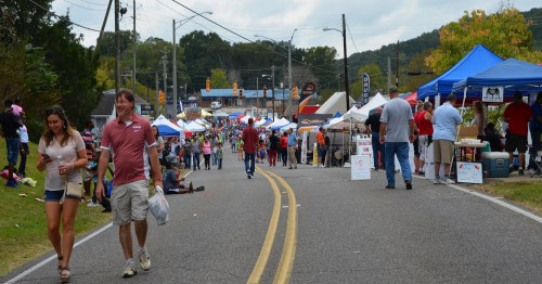 A lively street fair with vendors under blue tents, people walking, and colorful decorations in the background.