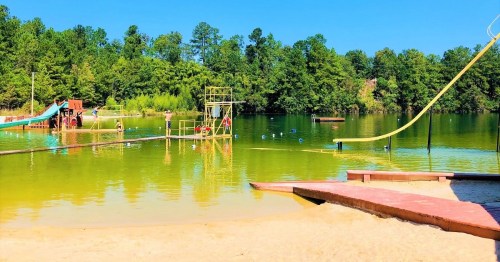 A sunny lakeside scene featuring a sandy beach, water slide, and playground equipment surrounded by trees.