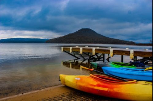 Colorful kayaks lined up on a sandy shore, with a wooden pier extending into a calm lake and a mountain in the background.