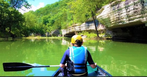 A person in a kayak paddles through a serene green river surrounded by lush trees and rocky cliffs.