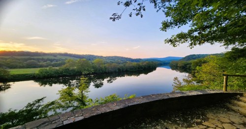 Scenic view of a calm river reflecting trees and hills at sunset, framed by lush greenery and a stone wall.
